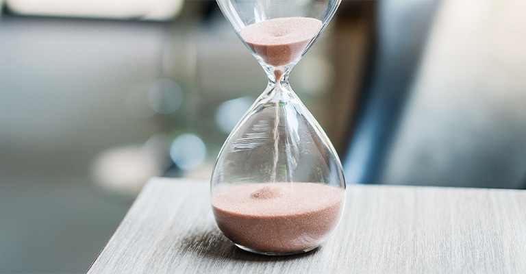 OffPageSEO_12 • Andrew Roche SEO A clear glass hourglass with pink sand sits on the edge of a light wooden surface, with sand visibly flowing from the top to the bottom chamber. The image symbolizes the passage of time, patience, or a countdown.