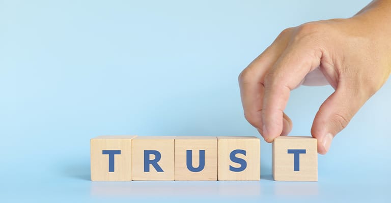 OffPageSEO_5 • Andrew Roche SEO A hand is placing the final wooden block to complete the word "TRUST" spelled out in bold blue letters on wooden cubes against a light blue background. The image conveys the concept of building or establishing trust.