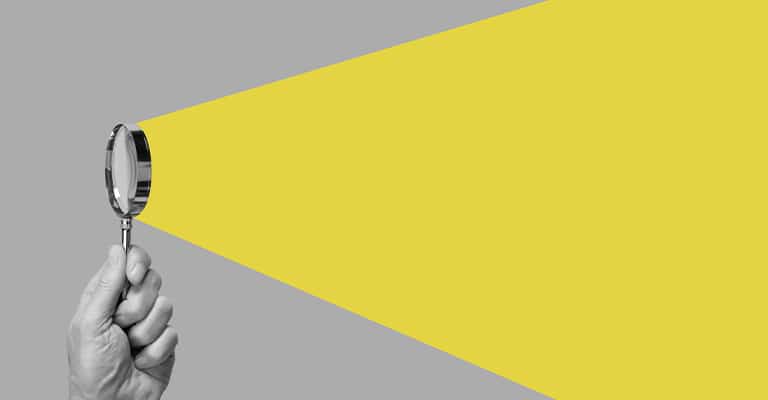 OffPageSEO_9 • Andrew Roche SEO A hand holds a magnifying glass against a gray background, with a bright yellow beam projecting from it like a flashlight. The image visually represents focused search, investigation, or discovery.